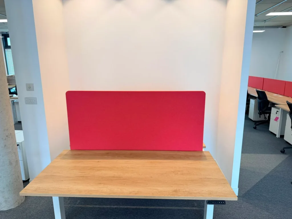 A minimalist wooden standing desk featuring a bold red privacy screen divider. This modern office workstation is situated in a bright, professional workspace, highlighting functional corporate design.