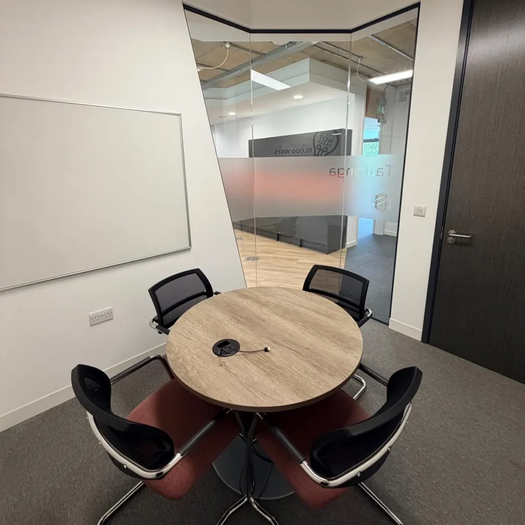 A compact, contemporary huddle room featuring a circular wooden table, ergonomic black chairs, and a wall-mounted whiteboard. This professional meeting space is enclosed by glass partitioning, offering a private yet open-feel environment for team collaboration.